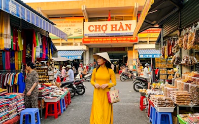 Woman in traditional dress at Chợ Hàn market, Da Nang, surrounded by colorful stalls and local goods.
