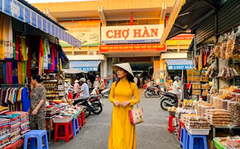 Woman in traditional dress at Chợ Hàn market, Da Nang, surrounded by colorful stalls and local goods.