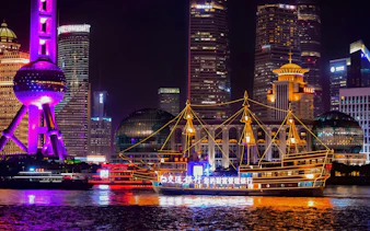 Traditional-style boat with yellow lights on Huangpu River, Shanghai, with city skyline at night.