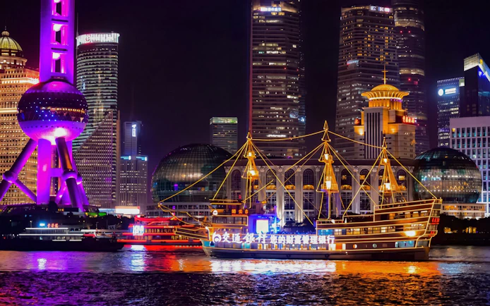 Traditional-style boat with yellow lights on Huangpu River, Shanghai, with city skyline at night.
