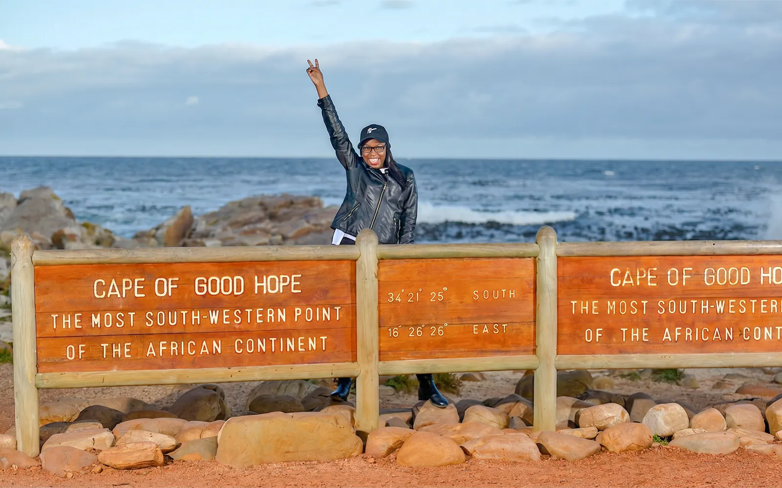 Person posing by Cape of Good Hope sign, Cape Town, with ocean in background.