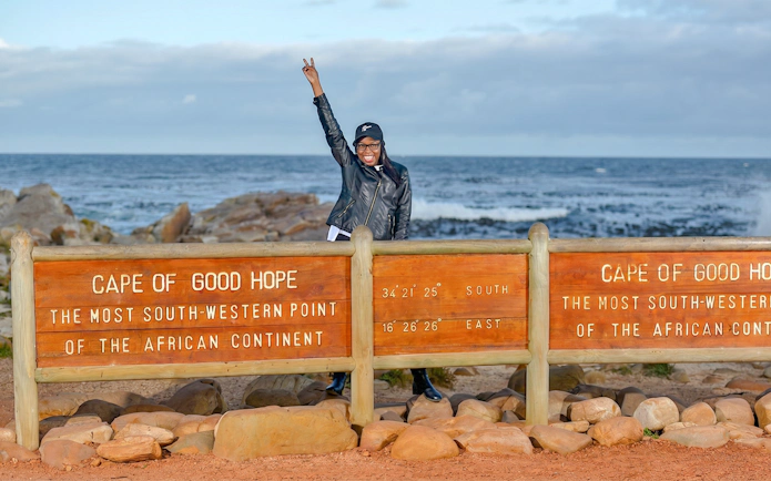 Person posing by Cape of Good Hope sign, Cape Town, with ocean in background.