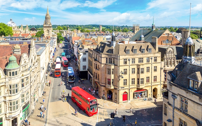 Buses on a busy street in Oxford, with historic buildings and spires in the background.