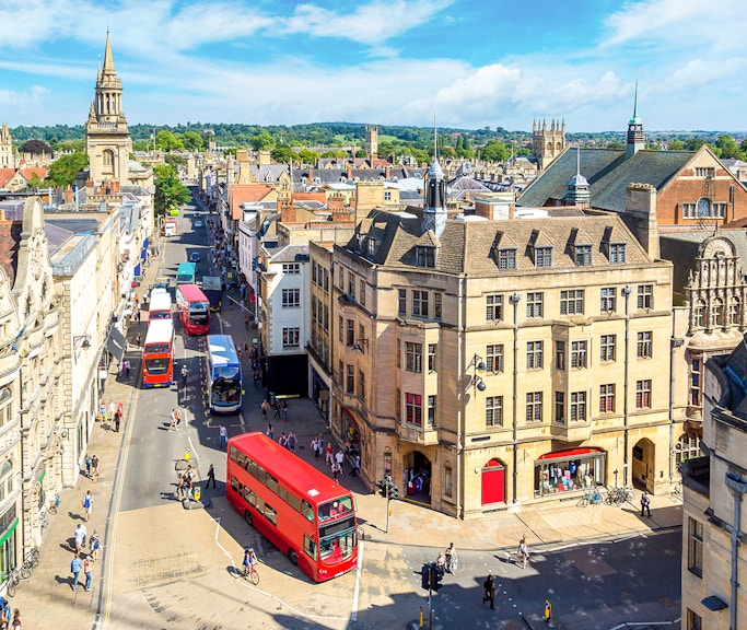 Buses on a busy street in Oxford, with historic buildings and spires in the background.