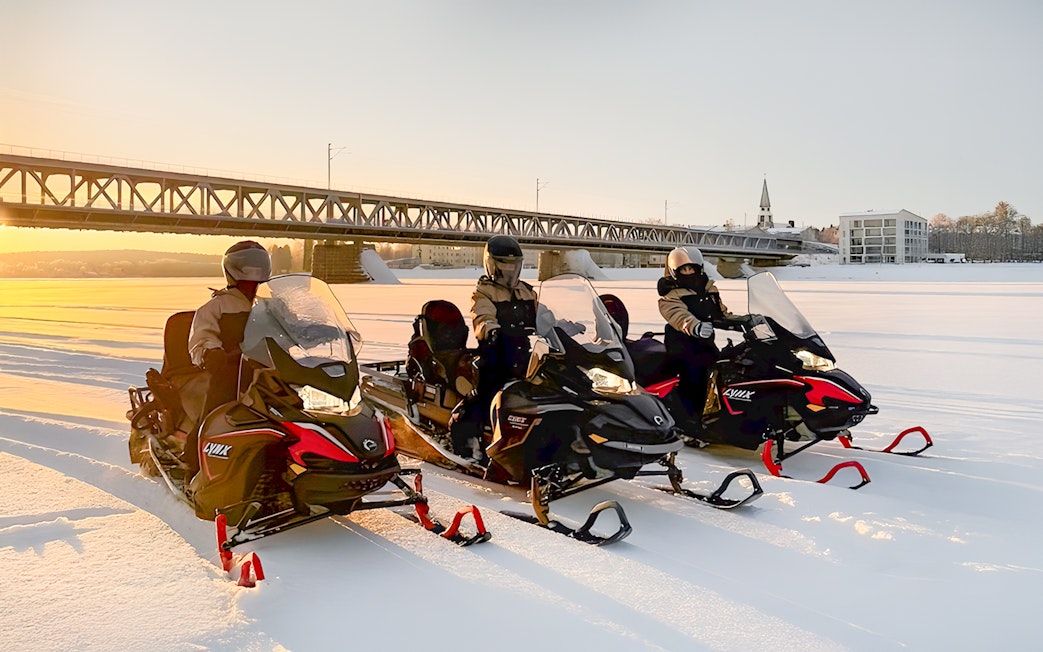 Snowmobilers crossing a frozen lake at sunset in Lapland, with a bridge in the background.