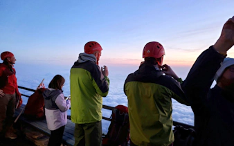 Climbers in helmets view sunrise above clouds on Mt. Fuji climbing tour.