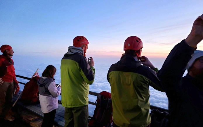 Climbers in helmets view sunrise above clouds on Mt. Fuji climbing tour.