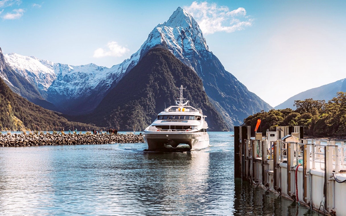 Cruise boat in Milford Sound with fjord views, lush mountains, and waterfalls, New Zealand.
