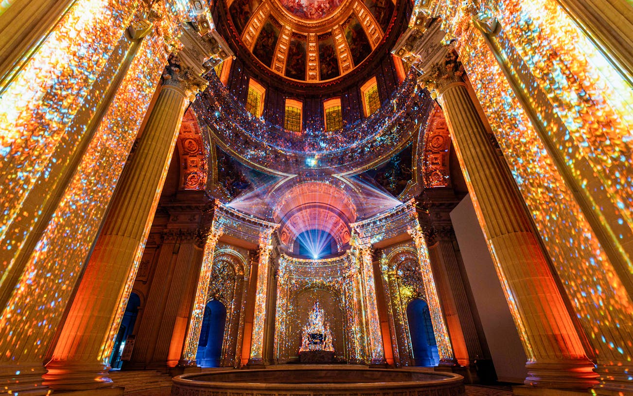 Immersive light display inside Invalides, Paris, showcasing vibrant projections on columns and ceiling.