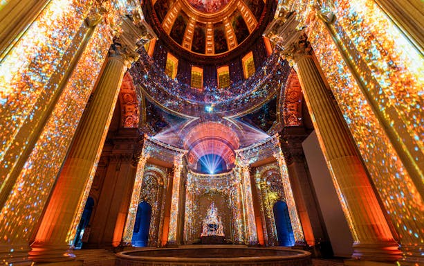 Immersive light display inside Invalides, Paris, showcasing vibrant projections on columns and ceiling.