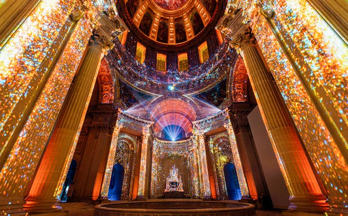 Immersive light display inside Invalides, Paris, showcasing vibrant projections on columns and ceiling.