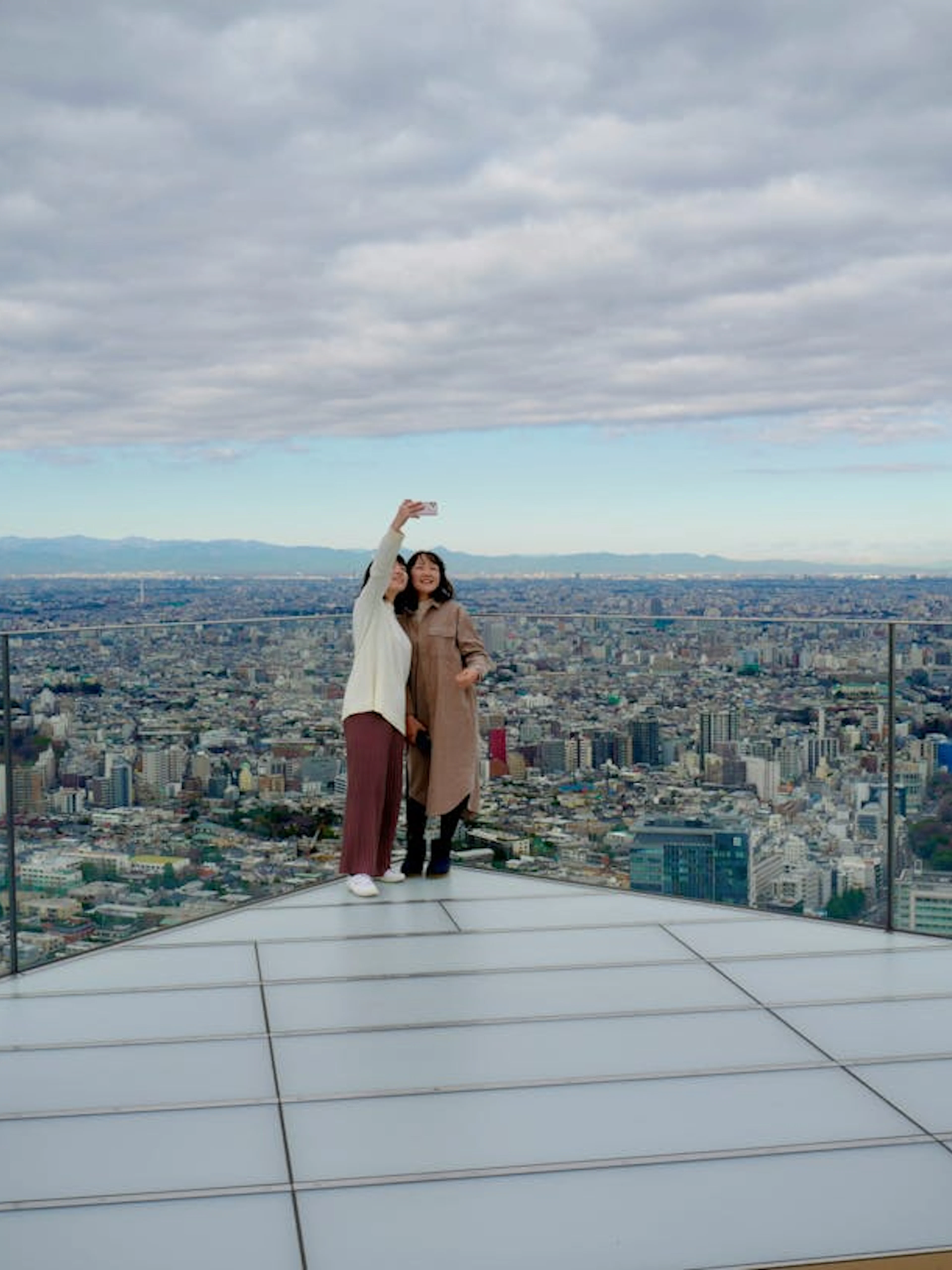 Visitors taking a selfie on Shibuya Sky observation deck with Tokyo cityscape in the background.