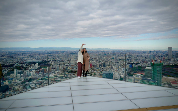 Visitors taking a selfie on Shibuya Sky observation deck with Tokyo cityscape in the background.