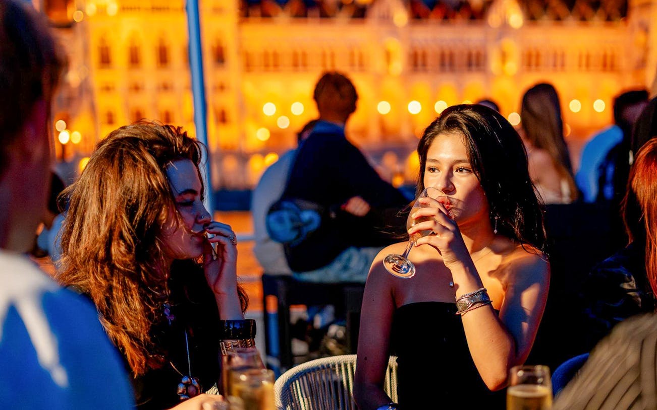 Guests enjoying drinks on Budapest Danube River nighttime cruise with illuminated cityscape.