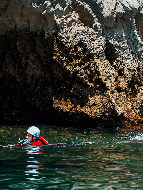 Adventurers coasteering near rocky cliffs in the ocean, wearing helmets and wetsuits.