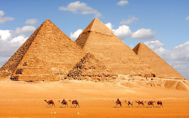 Camels walking in front of the Pyramids of Giza, Cairo, under a blue sky.