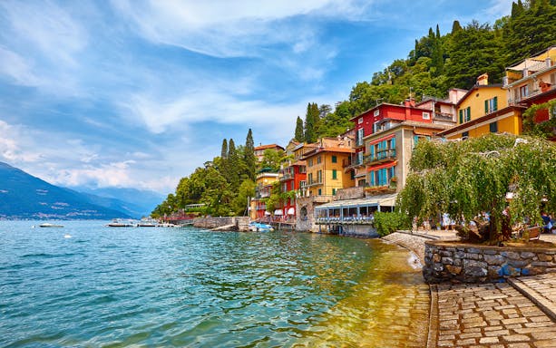Colorful buildings along Lake Como waterfront, Italy, with boats and lush greenery.