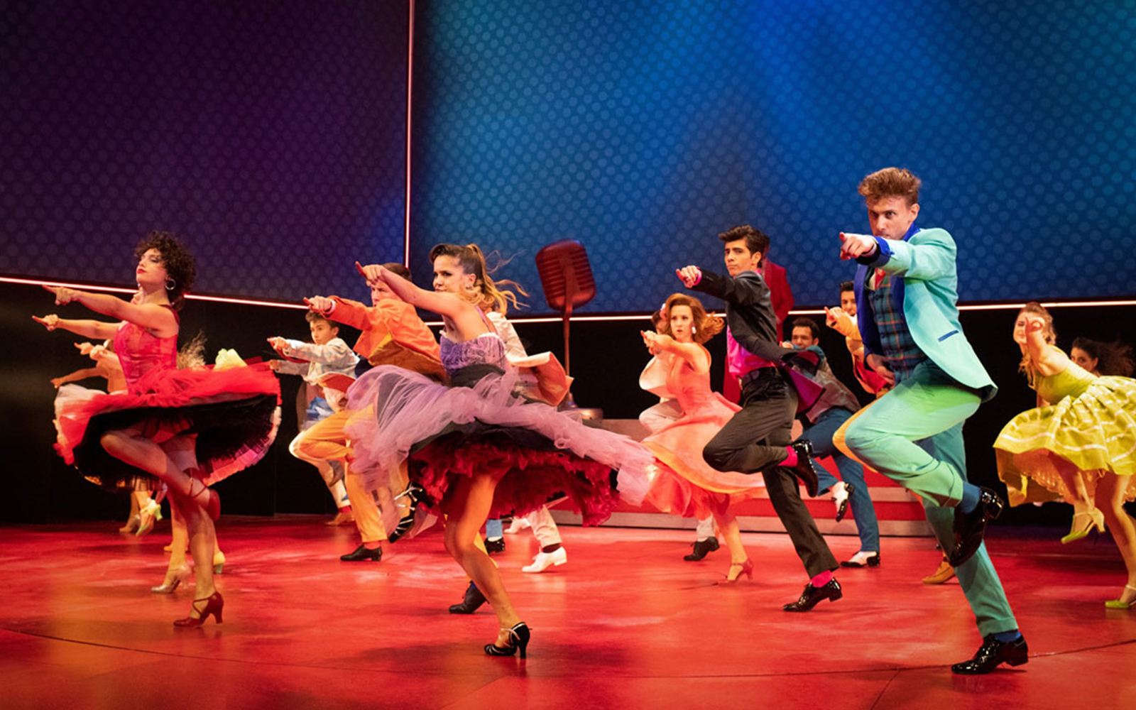 Dancers performing a lively routine in colorful costumes on stage during a Grease musical.
