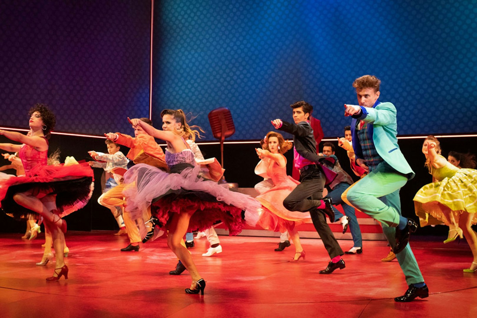 Dancers performing a lively routine in colorful costumes on stage during a Grease musical.