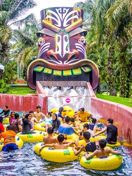 Water park visitors on yellow tubes at A'Famosa Malacca, Malaysia, near a colorful tribal mask slide.