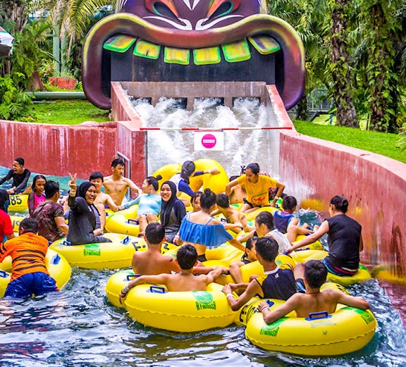 Water park visitors on yellow tubes at A'Famosa Malacca, Malaysia, near a colorful tribal mask slide.