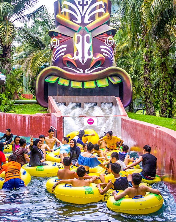 Water park visitors on yellow tubes at A'Famosa Malacca, Malaysia, near a colorful tribal mask slide.