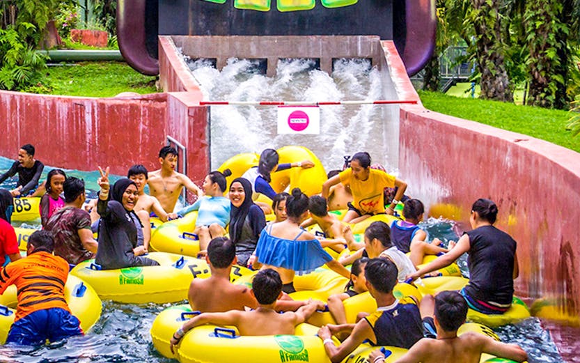 Water park visitors on yellow tubes at A'Famosa Malacca, Malaysia, near a colorful tribal mask slide.