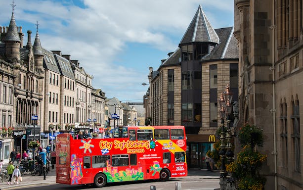 Red double-decker sightseeing bus on a street in Inverness, Scotland, with historic buildings.