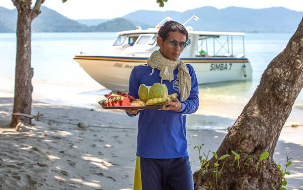Man holding a tray of tropical fruit on a beach with a boat in the background, Phi Phi Islands.