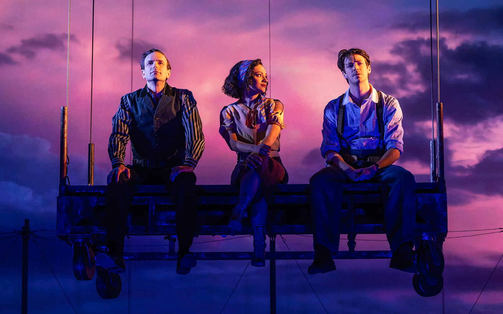 Three performers sit on a suspended platform against a dramatic purple sky in "Water for Elephants" stage scene.