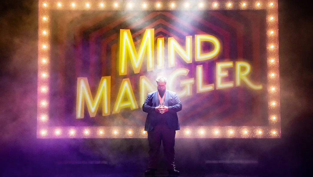 Mind Mangler performer on stage with illuminated sign in background.