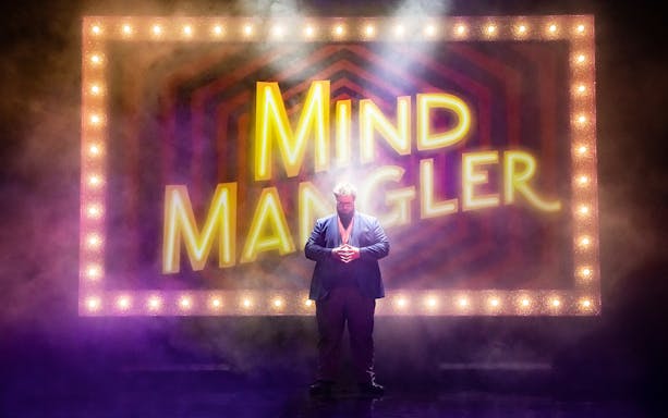 Mind Mangler performer on stage with illuminated sign in background.