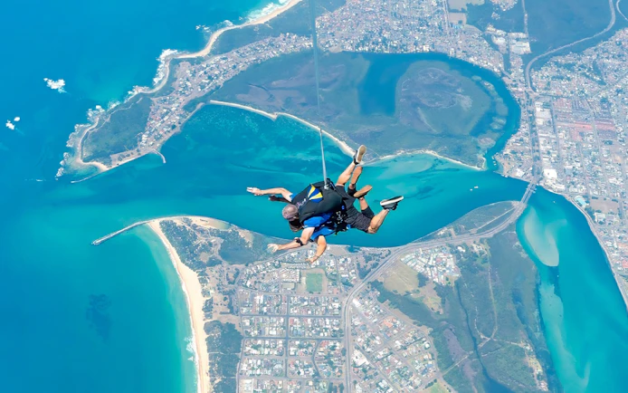 Tandem skydivers over Newcastle coastline, Australia, with ocean and cityscape below.