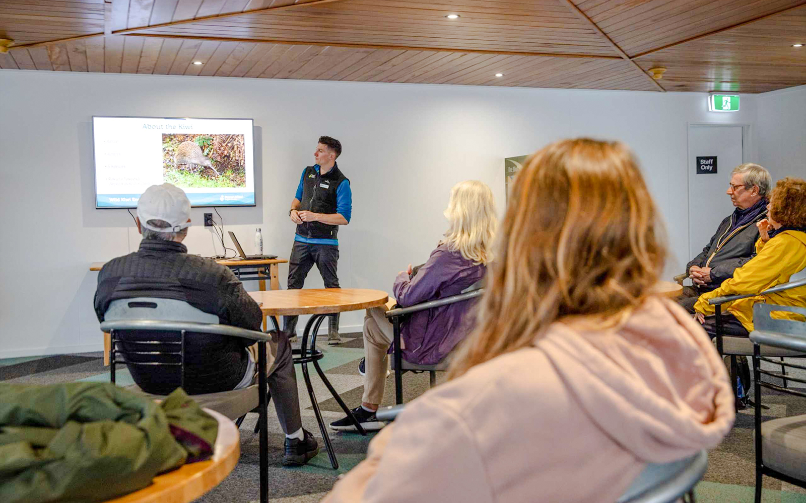 Guide presenting kiwi bird information to tourists in a room with a screen.