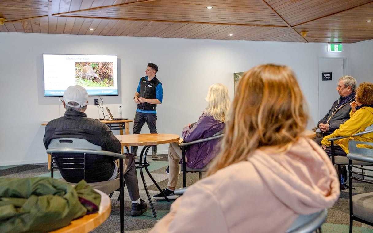 Guide presenting kiwi bird information to tourists in a room with a screen.