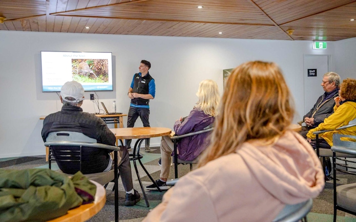 Guide presenting kiwi bird information to tourists in a room with a screen.