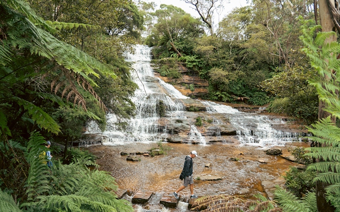 Man walking on rocks in front of Katoomba Cascades waterfall, surrounded by lush greenery.