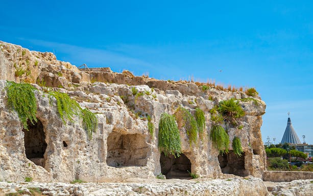 Ancient rock formations at Neapolis Archaeological Park, Syracuse, with a distant view of a modern structure.
