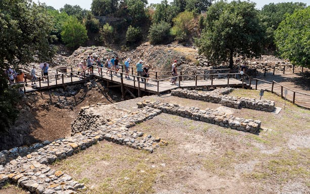Tourists exploring ancient ruins of Troy on a wooden walkway, surrounded by greenery.