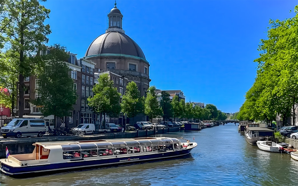 Canal cruise boat on Amsterdam canal with historic buildings and dome in the background.