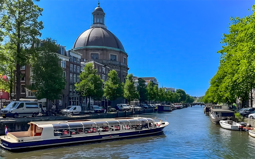Canal cruise boat on Amsterdam canal with historic buildings and dome in the background.