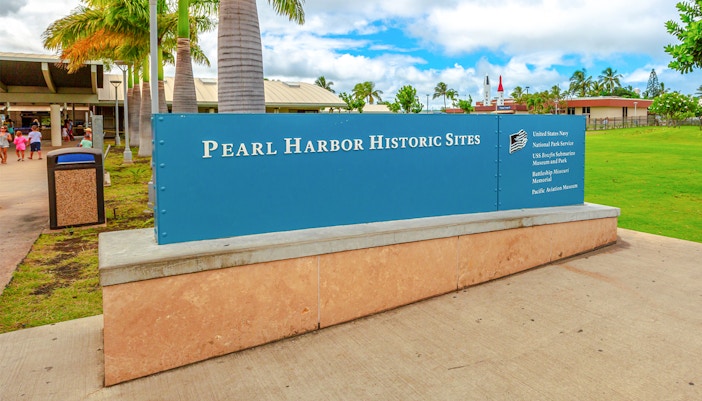 Pearl Harbor Visitor Center entrance sign with palm trees and buildings in the background.