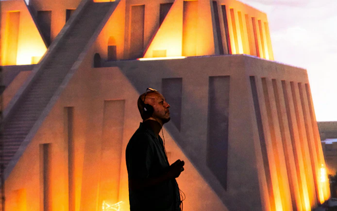 Man with headphones at London Timewalk Exhibition, illuminated ancient structure in background.