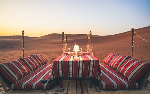 Private dinner setup with cushions and table in desert at sunset.