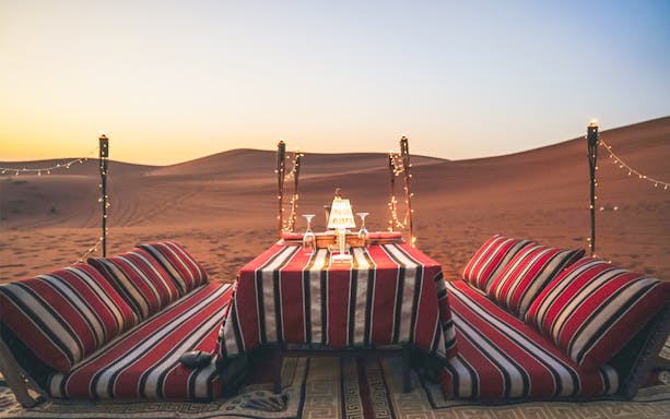 Private dinner setup with cushions and table in desert at sunset.