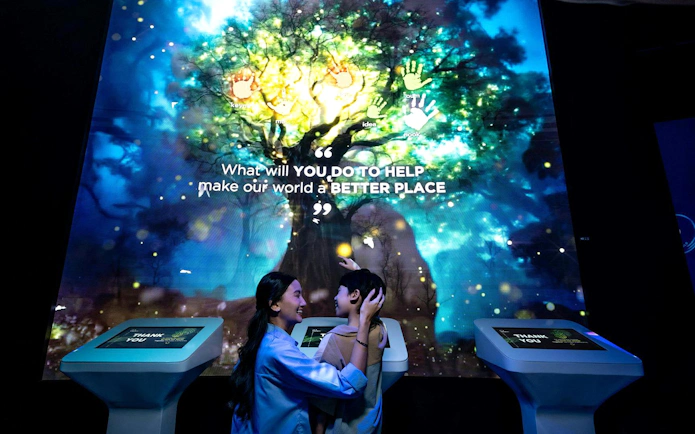 Interactive exhibit with a tree display encouraging a better world, featuring two people engaging with the installation.
