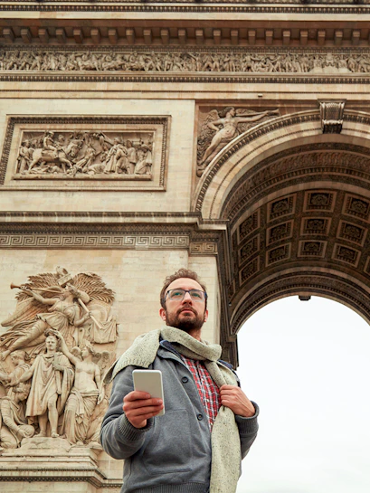 Man with smartphone in front of Arc de Triomphe, Paris, on audio-guided tour.