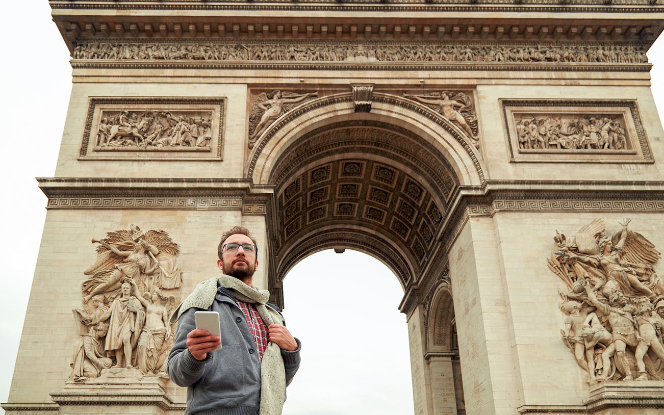 Man with smartphone in front of Arc de Triomphe, Paris, on audio-guided tour.