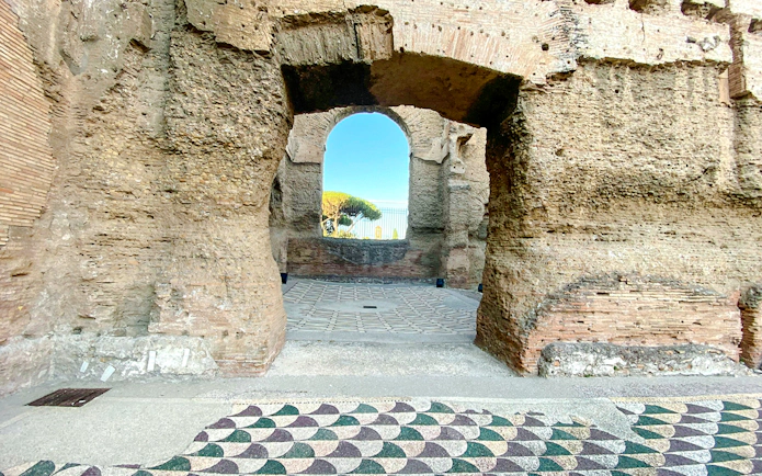 Stone archway at the Baths of Caracalla in Rome, Italy, with mosaic floor and distant trees.