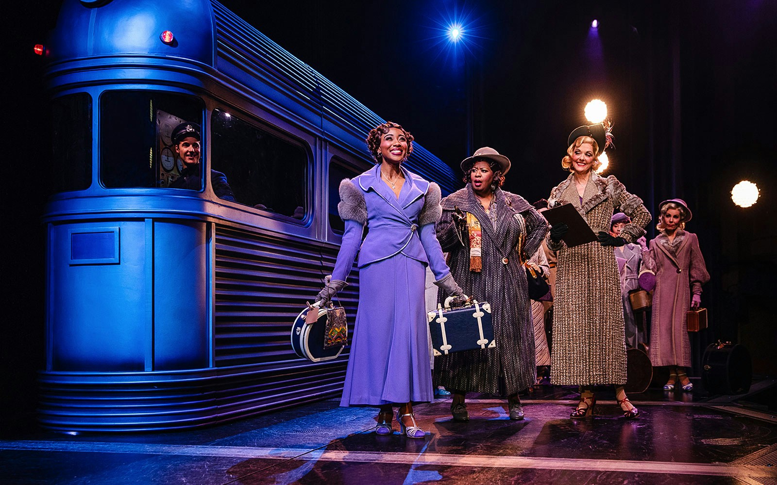 Women in vintage attire with suitcases beside a blue train on stage, "Some Like it Hot" musical.